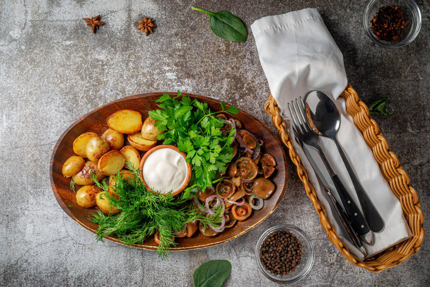 Serving a dish from a restaurant menu: country-style baked potatoes with pickled mushrooms and onions, cream sauce, dill and parsley greens on a plate against the background of a gray stone table - Фото, изображение