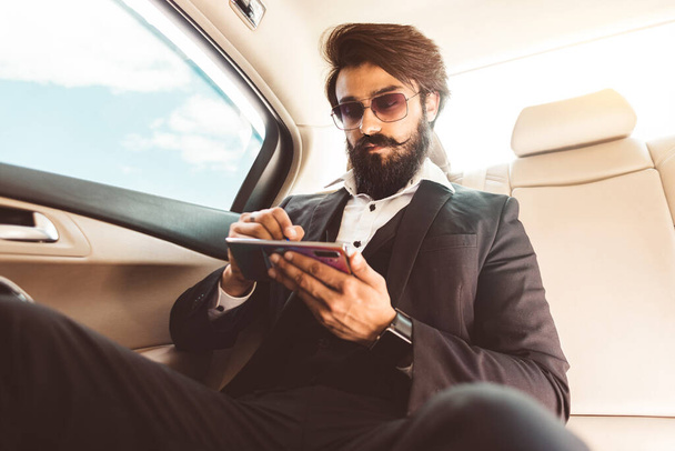 Stylish handsome Hindu businessman with glasses and a beard works in the backseat of the car and uses the phone. Safe and comfortable luxury travel - Foto, imagen