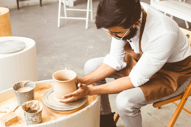 A young male potter is engaged in craft in his workshop on a potters wheel and makes a clay product - Foto, Imagem