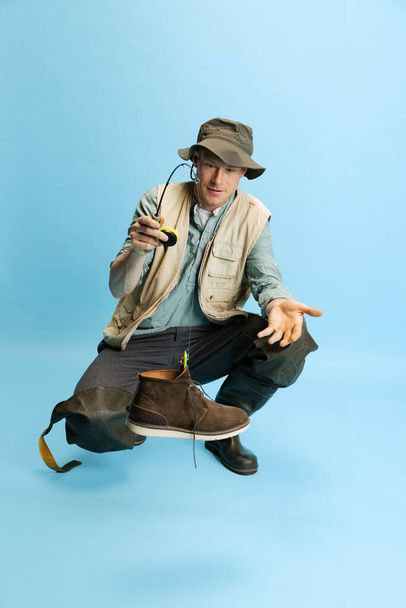 Funny young man, fisherman with fishing accessories wearing sport style clothes isolated over blue studio background - Zdjęcie, obraz