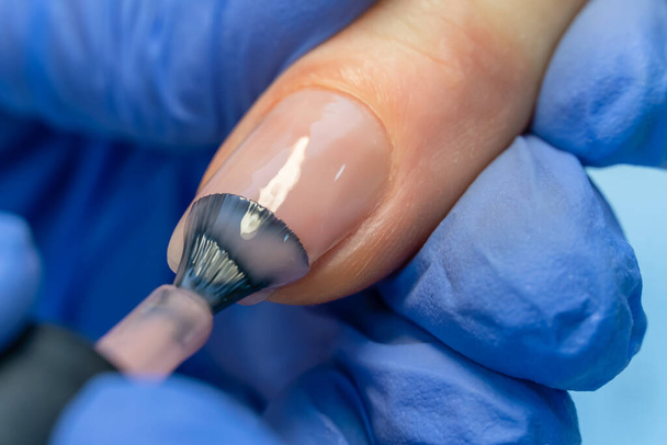 Close-up of a manicure master in blue gloves applies a layer of pink gel polish to the client's nail. Coating the nail plate with nail polish. Professional manicure in the salon close-up. Macro - Photo, Image