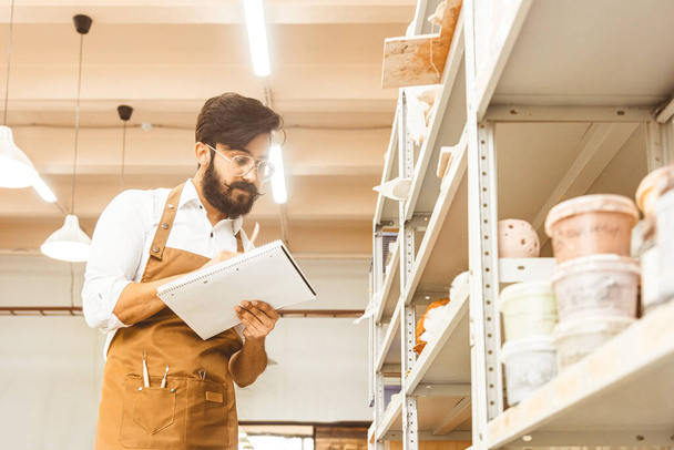 Young attractive businessman a potter with a beard and mustache works in his workshop. Keeps records and transcribes into a notebook inspecting shelves with products, conducts an inventory - Фото, изображение