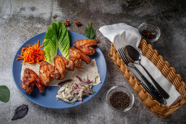 Serving a dish from the restaurant menu. Chicken wings, grilled nuggets with grated carrots and herbs on a plate against a gray stone table, delicious kebab - Fotó, kép