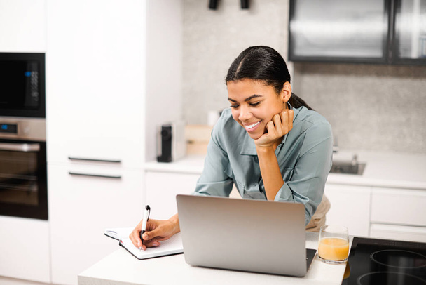 Θετική έξυπνη νεαρή μικτή φυλή γυναίκα φοιτητής χαμογελώντας, ενώ στέκεται στο τραπέζι με φορητό υπολογιστή στην κουζίνα, μελετώντας από το σπίτι και κρατώντας σημειώσεις για το σημειωματάριο, βλέποντας ένα online webinar - Φωτογραφία, εικόνα