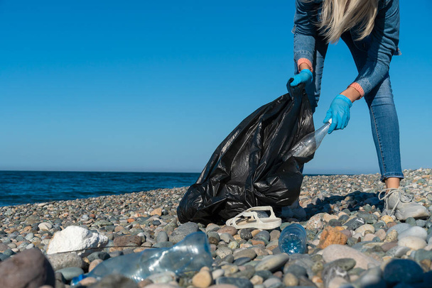 Genç bir kadın deniz kıyısındaki çakıl taşı plajında siyah bir çöp torbasında plastik topluyor. Kullanılmış kirli plastik şişeler. Karadeniz kıyısındaki çevre kirliliği - Fotoğraf, Görsel