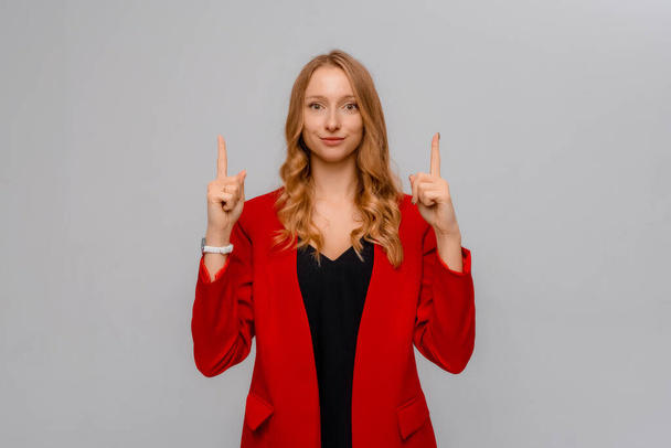 Retrato de una joven rubia sonriente apuntando con los dedos hacia arriba, mostrando una gran oferta promocional, venta en tienda de publicidad, de pie en chaqueta roja sobre fondo gris - Foto, imagen