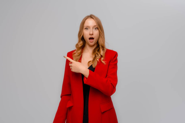 Hey look there. Excited blonde young woman shouts and looks with happy disbelief, pointing finger to the left, showing advertisement, stands in red blazer against gray background - Photo, image