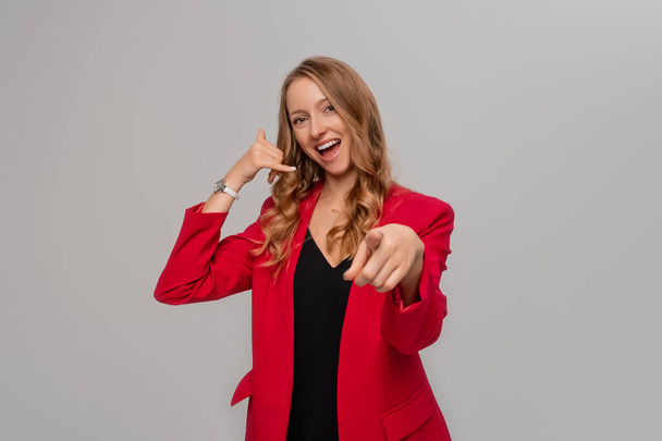 Happy beautiful young blonde woman pointing finger at camera, showing mobile phone call gesture, inviting you, standing in red blazer against gray background - Zdjęcie, obraz