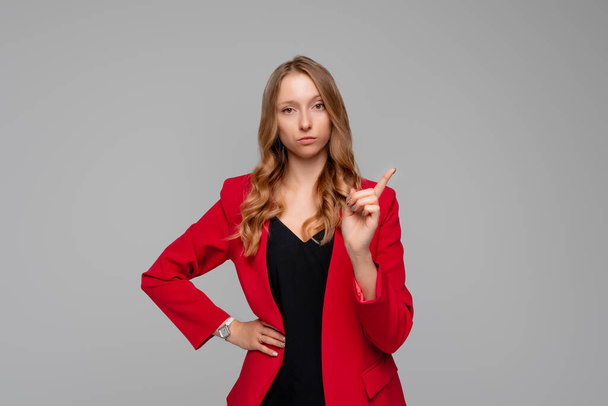 Σοβαρή γυναίκα επιχειρηματίας με ξανθά μαλλιά σε κόκκινα και μαύρα ρούχα δείχνοντας το δάχτυλο προς τα πάνω, κατσαδιάζοντας ή λέγοντας off κάποιον, λέει: Προσοχή παρακαλώ, κρατώντας το χέρι ισχίου αυτοπεποίθηση - Φωτογραφία, εικόνα