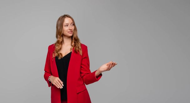 Confident successful blonde young woman pointing hand at at something interesting and showing advertisement, promo offer, standing in red blazer against gray background - Photo, image