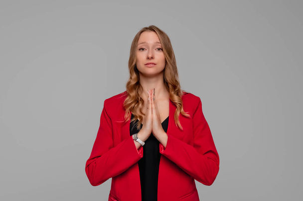 Hope Concept. Hopeful pretty Young Woman Holds Hands In Prayer Gesture, standing in red blazer against gray background. May my dreams come true - Фото, изображение