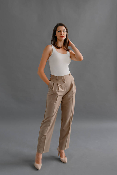 Full length portrait of happy businesswoman looking confident at camera, dressed in white t shirt with short sleeves and beige pants, posing in studio over gray background - Photo, Image