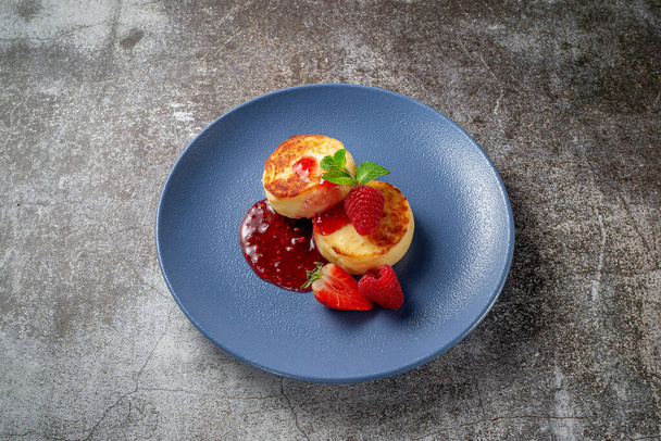 Fried pancakes with strawberry jam and raspberries and mint in a blue plate against a gray stone table. A sweet breakfast in the morning  - Photo, Image