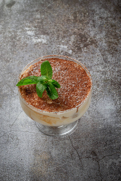 Classic tiramisu dessert with mint leaves in a glass, with cocoa and chocolate in the background of a gray stone table  - Fotó, kép