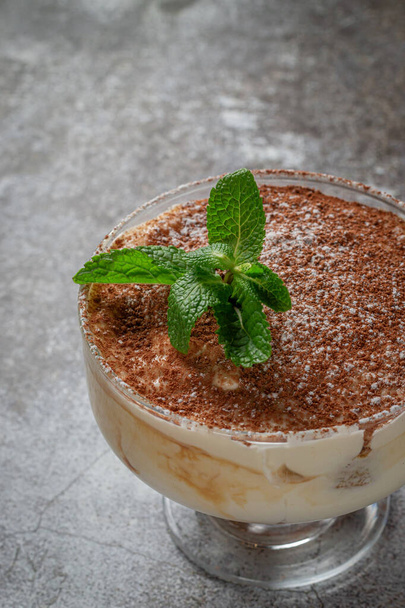 Classic tiramisu dessert with mint leaves in a glass, with cocoa and chocolate in the background of a gray stone table  - Foto, Imagen