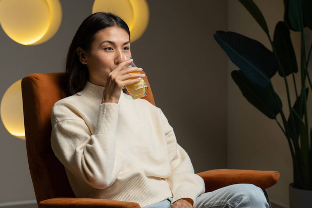 Asian woman drinks orange juice from a glass while sitting on the couch and relaxing - Foto, Bild