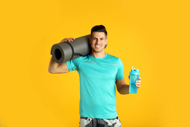 Handsome man with yoga mat and shaker on yellow background - Fotografie, Obrázek