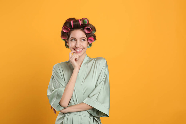 Happy young woman in silk bathrobe with hair curlers on yellow background. Space for text - Foto, imagen