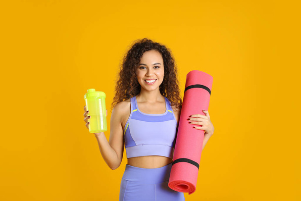 Beautiful African American woman with yoga mat and shaker on yellow background - Fotoğraf, Görsel