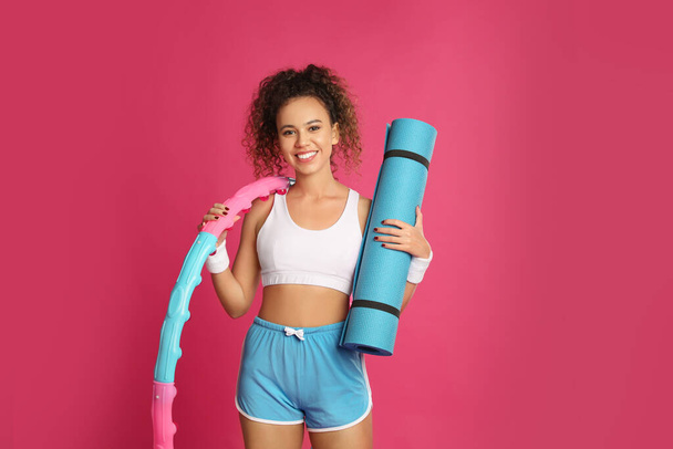 Beautiful African American woman with yoga mat and hula hoop on pink background - Valokuva, kuva