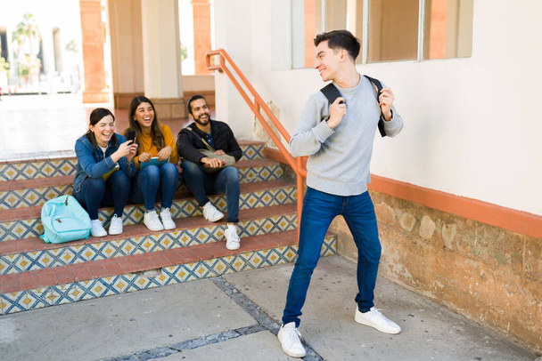 Διασκεδαστικός μαθητής κουβαλάει ένα σακίδιο και αστειεύεται ενώ οι φίλοι του γελούν και ηχογραφούν ένα βίντεο στο smartphone για να το ποστάρουν στα μέσα κοινωνικής δικτύωσης  - Φωτογραφία, εικόνα