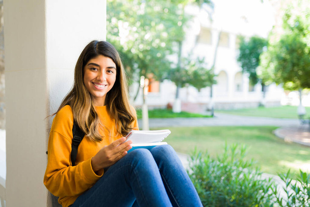 Smart college girl making eye contact and smiling while studying for her exam before class - Photo, Image