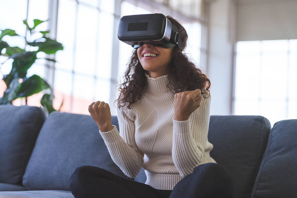 Young African American woman wearing virtual reality glasses is playing vr video games on sofa at home. Stay at home and self-quarantine in spread of coronavirus covid-19 - Foto, Bild