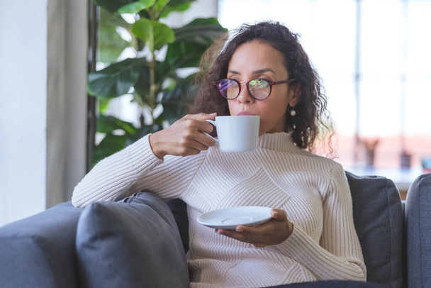 Retrato de una hermosa joven afroamericana con anteojos y café relajante y bebiendo en el sofá de vacaciones en casa. mujeres de negocios y autónomos que trabajan desde casa para propagar coronavirus covid-19 - Foto, imagen