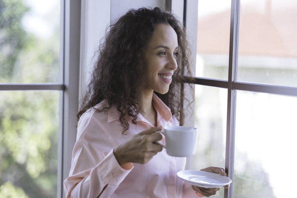 портрет красива молода латинка, що тримає чашку гарячої кави або гарячого чаю, посміхається, і дивиться на вікна з ранковим світлом. бізнес-леді розслабляється перед роботою. Щаслива жінка залишається вдома
. - Фото, зображення