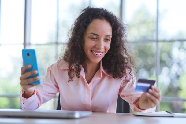 Молода латинка посміхається і використовує смартфон для онлайн-покупки. Бізнес-леді і позаштатна робота з дому і тримання кредитної картки для електронних платежів. щасливі послуги покупця в інтернет-магазині
. - Фото, зображення