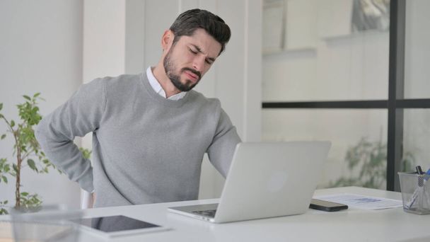 Giovane uomo che ha mal di schiena durante l'utilizzo del computer portatile in ufficio - Foto, immagini