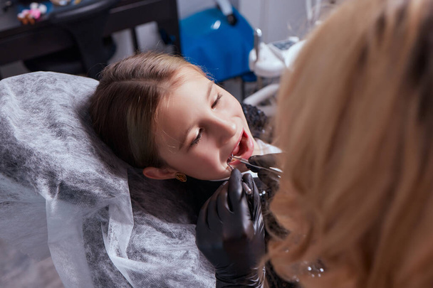 Caucasian young child girl sitting in medical chair while dentist fixing teeth at dental clinic using dental tools instruments, close-up photo of face. professional medical specialist. - Foto, Imagen