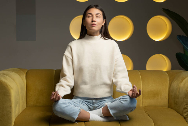 Time for yoga classes. Attractive young asian woman trains and sits in a yoga lotus position on the couch - Photo, image