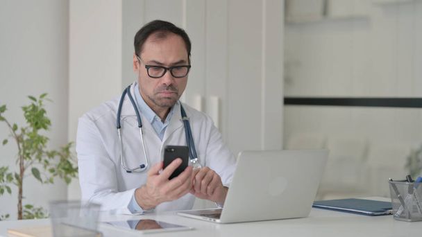 Middeleeuws arts met behulp van Smartphone tijdens het werken op laptop in de kliniek - Foto, afbeelding