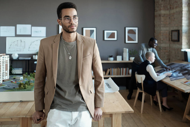 Молодий серйозний бізнесмен в розумному повсякденному одязі стоїть за дерев'яним столом
 - Фото, зображення