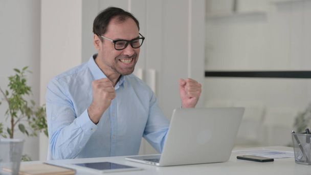Людина середнього віку Святкування під час використання ноутбука в офісі на лавці
 - Фото, зображення