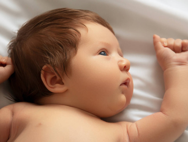 Ładny nowonarodzony chłopiec, portret z bliska. Leży w białym łóżku. Cute noworodka 1 miesiąc życia na białym prześcieradle w pieluszce. Pielęgnacja noworodków, kolka, zęby, noworodek - Zdjęcie, obraz