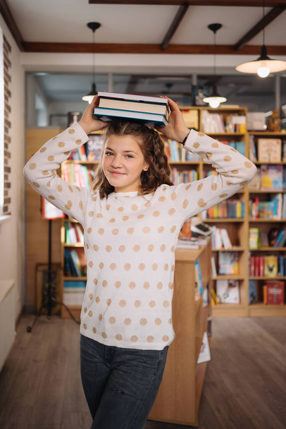 Beautiful girl is holding stack of books while standing among books in the bookshop - Photo, Image