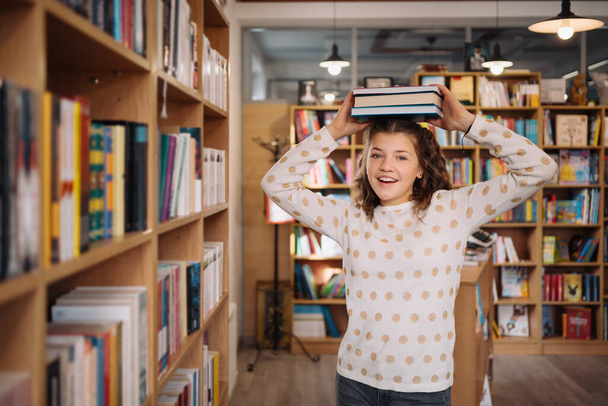 Beautiful girl is holding stack of books while standing among books in the bookshop - Photo, Image