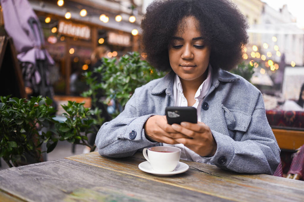 Street style portrait of attractive young happy African American woman with afro hair have video chat with smartphone outdoors in sidewalk cafe. Excited smiling female blogger broadcasts live stream. - Foto, Imagen