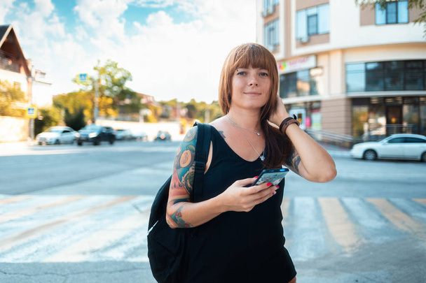 Portrait of a young smiling woman with a sleeve tattoo standing on the street and holding a smartphone. Street in the background. The concept of freedom and travel. - Foto, Bild