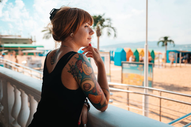 Portrait of a young smiling woman with a tattoo on her arm, looking at the sea. There is a beach in the background. The concept of summer vacation. - Photo, Image