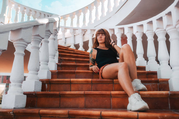 Beautiful young smiling woman with a sleeve tattoo sitting on the steps of a stone staircase. Bottom view. The concept of freedom and travel. - Foto, imagen