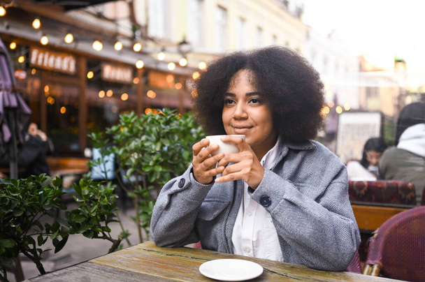 Fashion street style portrait of attractive young natural beauty African American woman with afro hair in tweed jacket posing outdoors in sidewalk cafe. Happy lady with coffee cup in big city. - Photo, image