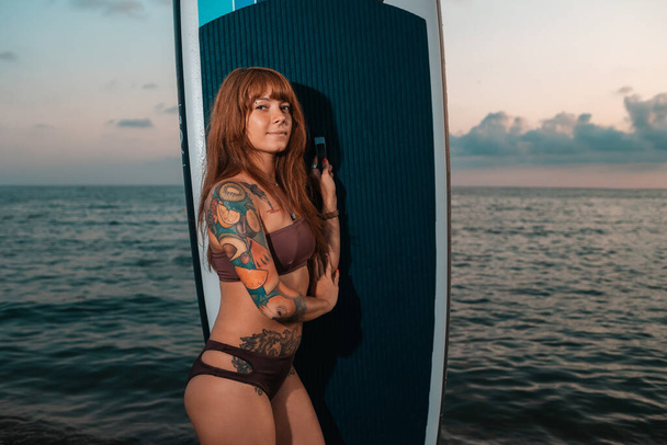 Portrait of tanned young woman with a tattoo poses standing with a sup board. The concept of active lifestyle and surfing. - Photo, image
