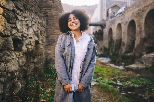 Fashion street style portrait of attractive young natural beauty African American woman with afro hair in blue coat posing outdoors. Happy tourist laughing walks through ancient sights fool around. - Фото, зображення