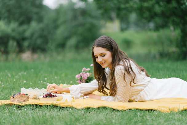 Mosolygó kaukázusi tini lány ül a szabadban. Kreatív művész fiatal nő visel alkalmi ruhák, amelynek piknik gyümölcsökkel a nyári napon. - Fotó, kép