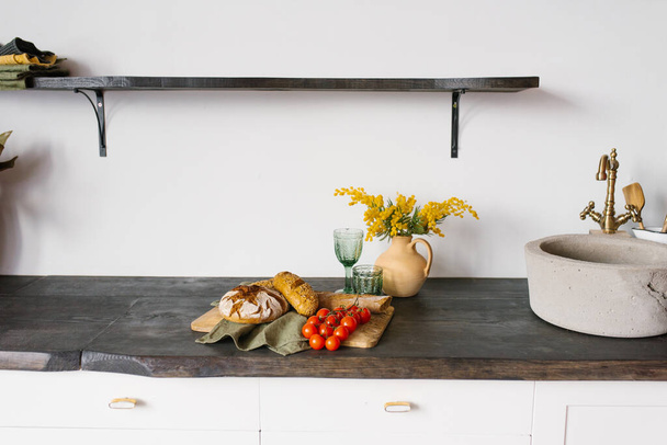 Cucina luminosa e moderna con lavello in pietra. Pani di pane fresco fatto in casa e un rametto di pomodoro su un tagliere, mimosa in un vaso di argilla. arredamento nella cucina scandinava - Foto, immagini
