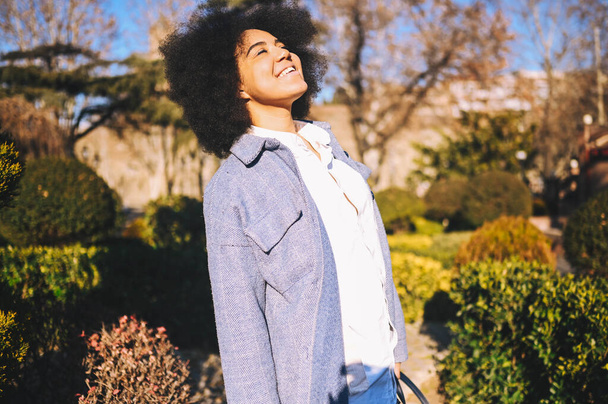 Close up Fashion street style portrait of attractive young natural beauty African American woman with afro hair in blue coat posing walking outdoors in sunny day. Happy lady with perfect teeth smile. - Fotoğraf, Görsel