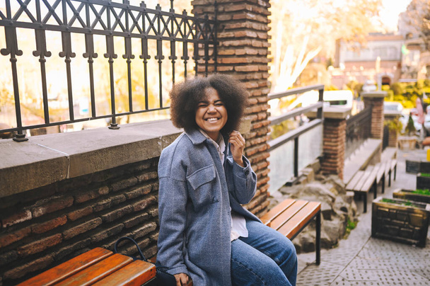 Fashion street style portrait of attractive young natural beauty African American woman with afro hair in blue coat posing outdoors. Happy tourist laughing walks through ancient sights fool around. - Fotoğraf, Görsel